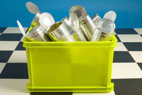 A lime green plastic bin positioned on a surface with a black and white checkered pattern, filled with a collection of empty, silver tin cans that are stacked and leaning against each other. The cans have a reflective metallic finish and display visible ridges and open tops, with some appearing slightly dented or used. The background features a solid blue wall, creating a contrasting backdrop for the bin and contents. This image exemplifies a typical waste collection scene, relevant to rubbish removal services in the Mortlake area, where Garden Clearance Mortlake offers garden waste and general rubbish disposal solutions. The scene captures the practical aspect of waste collection, with an emphasis on metal containers and clutter, suitable for illustrating content related to waste removal, recycling, and clearance activities commonly undertaken in local residential or commercial settings within the postcode area of SW14 or nearby South West London neighborhoods.