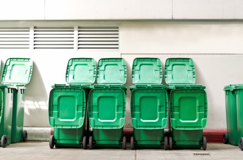 A row of ten green plastic wheelie bins with closed lids, arranged in two groups of five, positioned on a paved surface against a white wall with ventilation slots. The bins, used for rubbish and waste collection, are aligned neatly with their wheels and handles visible, and are situated outdoors in a residential or commercial area near a mortar-red painted curb. Natural daylight illuminates the scene, casting soft shadows. The location suggests a typical rubbish disposal area, possibly adjacent to a property in Mortlake, as indicated by the postcode inference. This setup aligns with common waste management practices in the UK, with local companies like Garden Clearance Mortlake facilitating waste and rubbish removal services in the area.