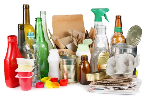 A collection of various discarded household items including glass bottles in green, brown, and clear colors, some with labels, and plastic bottles in red, green, and transparent with caps. There are metal cans and foil containers stacked among paper and cardboard waste, such as crumpled boxes and shredded paper. A plastic spray bottle with a green trigger is visible among the rubbish, alongside crumpled tissues or paper towels, and a few fabric-like materials. The items are arranged on a surface, possibly in a domestic or garden setting, with some items leaning on others. The image emphasizes different materials commonly found as garden or household waste, reflecting types of rubbish that Garden Clearance Mortlake may remove in the local Mortlake area. The lighting appears natural, with no distinct shadows, highlighting the variety of textures from smooth glass to crinkled paper and soft tissue. This detailed visual captures the type of waste that can be collected and disposed of through professional rubbish removal services in the Mortlake region of southwest London, incorporating elements relevant to local property types and waste streams.