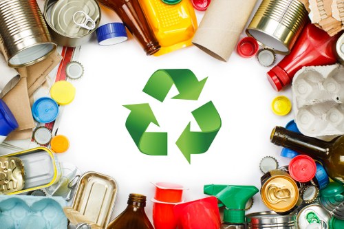 A collection of various household waste materials arranged in a circular formation around a prominent green recycling symbol. The waste includes aluminium cans, glass bottles in brown, green, and clear, plastic bottles in yellow, red, and blue, along with plastic containers, foil wrappers, and some cardboard pieces. The cans and bottles display metallic, glossy, and textured surfaces, while the cardboard has a matte, rough finish. The objects are positioned on a clean, white background, with some overlapping and oriented in different directions, reflecting the typical contents handled during rubbish removal services in the Mortlake area. The recycling symbol in the center signifies the importance of waste sorting and environmental responsibility, relevant to the services offered by Garden Clearance Mortlake for garden waste and rubbish removal in this locality.