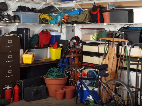 A densely packed garage interior displaying various household and outdoor equipment. In the foreground, there is a blue bicycle leaning against a wooden shelving unit, along with several empty terracotta plant pots stacked on the concrete floor. To the left, a black storage drawer unit and a black box are visible. Multiple plastic storage containers in blue, grey, and black are arranged on metal shelving units that extend across the background, some with lids and others open, containing tools, gardening supplies, and miscellaneous items. A yellow bucket and a red carrier are placed on the ground, alongside a coiled green garden hose and a small speaker. The upper shelves hold additional items, including coiled cords, gloves, and small boxes. The scene suggests a typical suburban garden clearance storage space, potentially located in Mortlake, with various tools and containers stored for outdoor maintenance and rubbish removal services by Garden Clearance Mortlake.