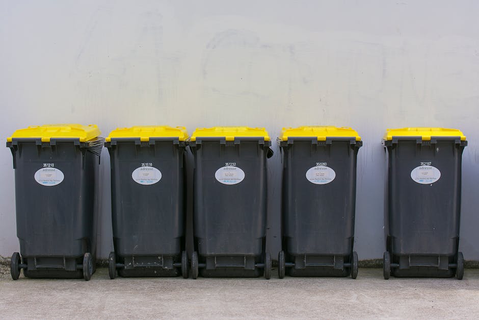 A worker wearing a yellow and red high-visibility vest is standing in front of a large red rubbish collection truck parked on the side of a street. The worker appears to be operating the waste compact