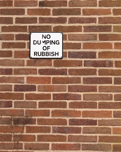 A rectangular white metal sign affixed to a red brick wall features black uppercase text that reads 'NO DUMPING OF RUBBISH'. The sign is positioned centrally on the wall, which has a uniformly laid br